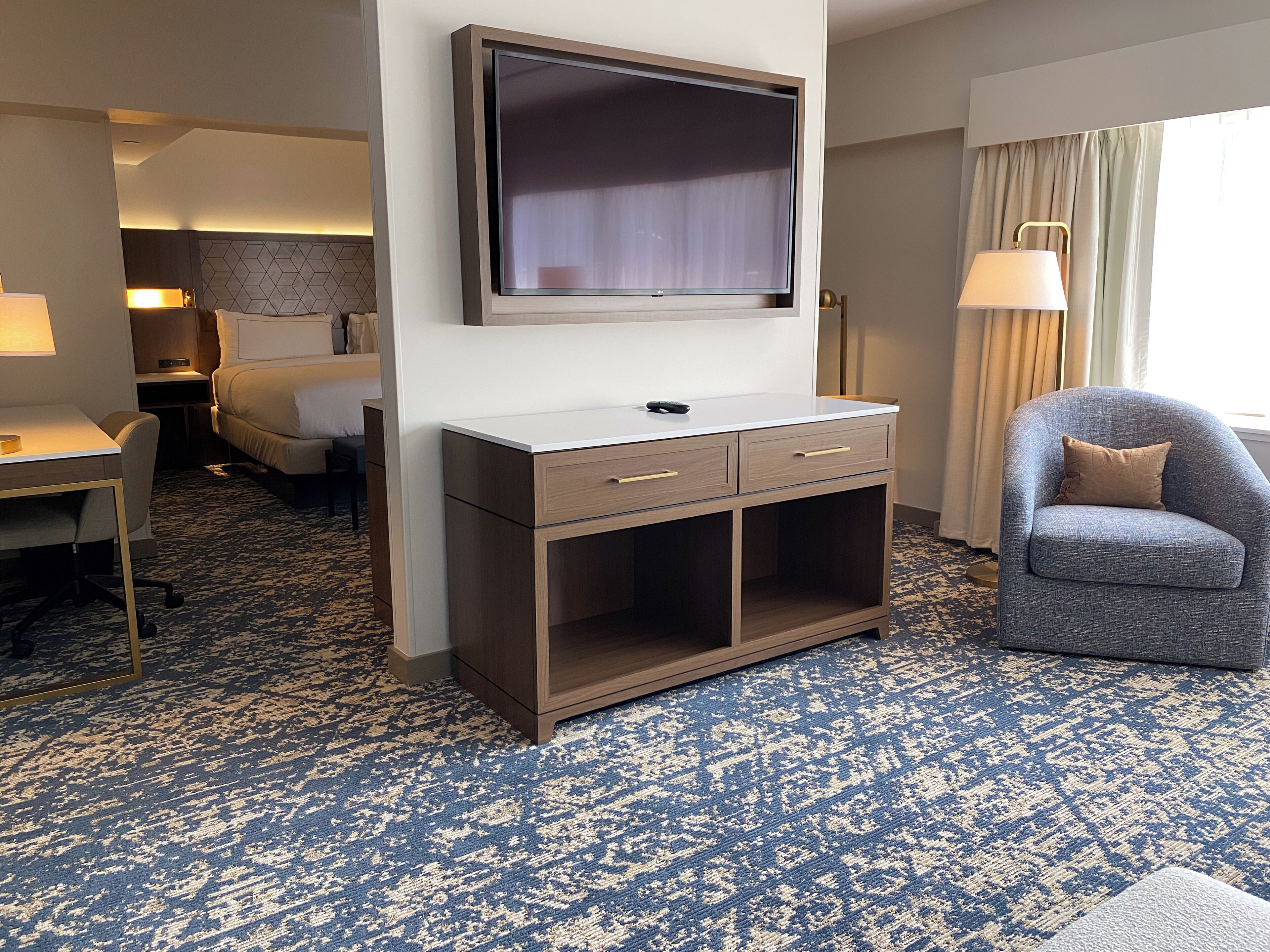 A hotel room with a flat-screen TV mounted on the wall, a desk with a lamp, a chair, and a bed.