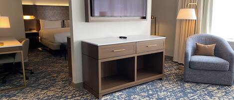 A hotel room with a flat-screen TV mounted on the wall, a desk with a lamp, a chair, and a bed.