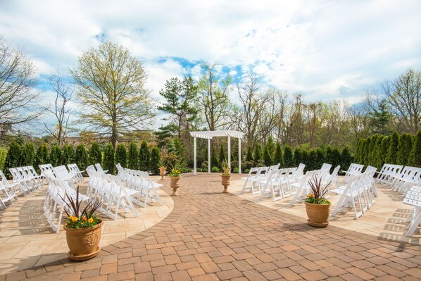 An outdoor wedding venue with white chairs arranged in rows, a central white gazebo, and potted plants.