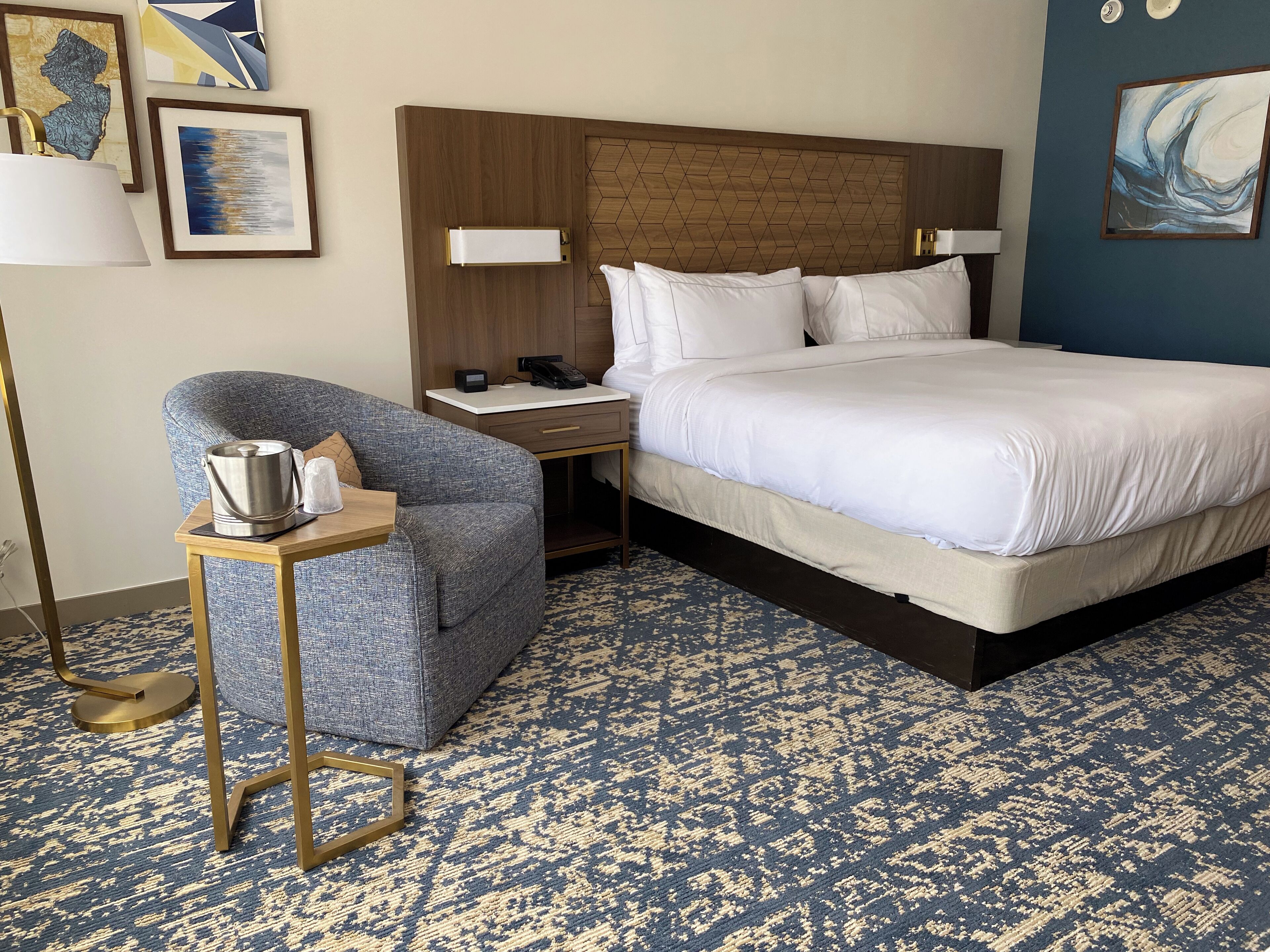 A hotel room with a large bed, a seating area with a chair, a bedside table with a lamp, and artwork on the walls.