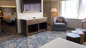 A hotel room with a large flat-screen TV mounted on the wall, a grey armchair, a small wooden desk, and a patterned carpet.
