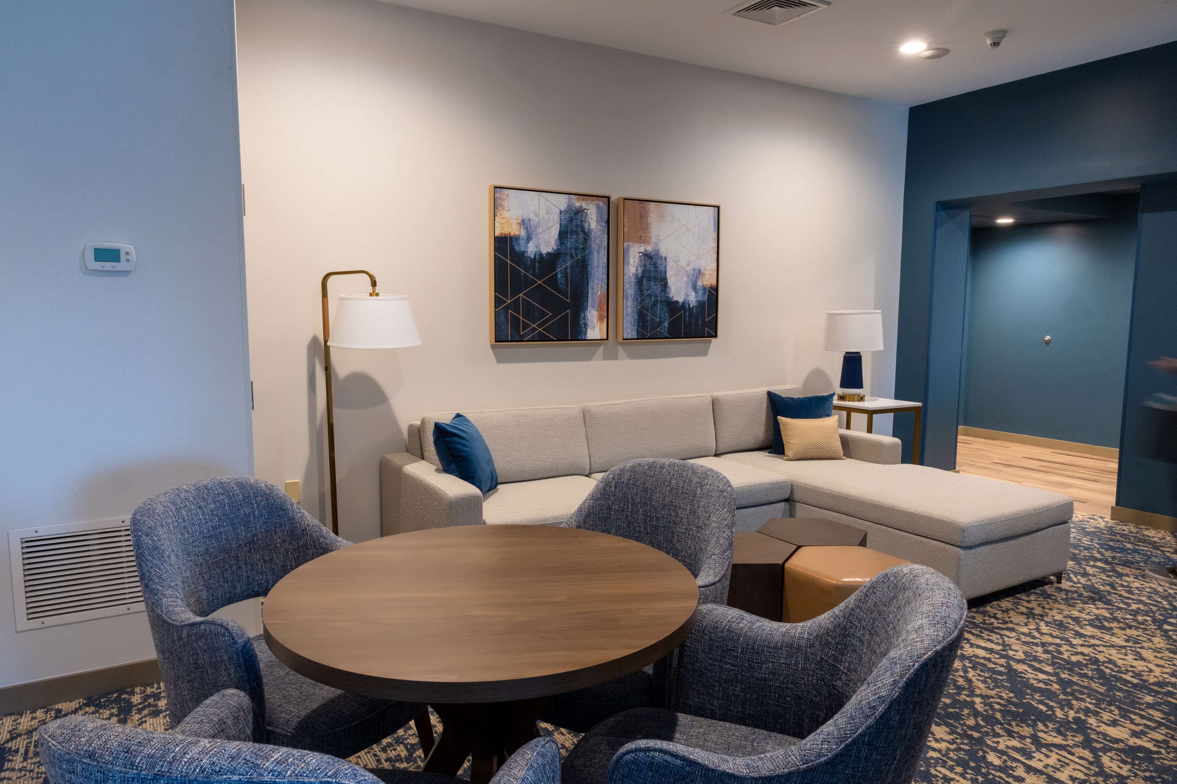 A modern hotel room with a sectional sofa, round dining table, and two wall-mounted paintings.