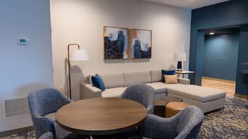 A modern hotel room with a sectional sofa, round dining table, and two wall-mounted paintings.