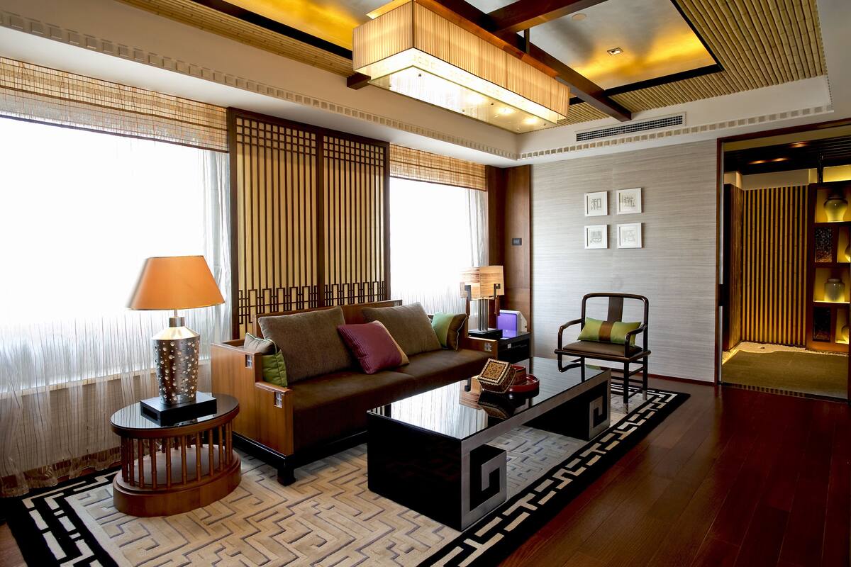 A modern living room with a sofa, coffee table, and lamps. There is a window with sheer curtains and a view of the outside.