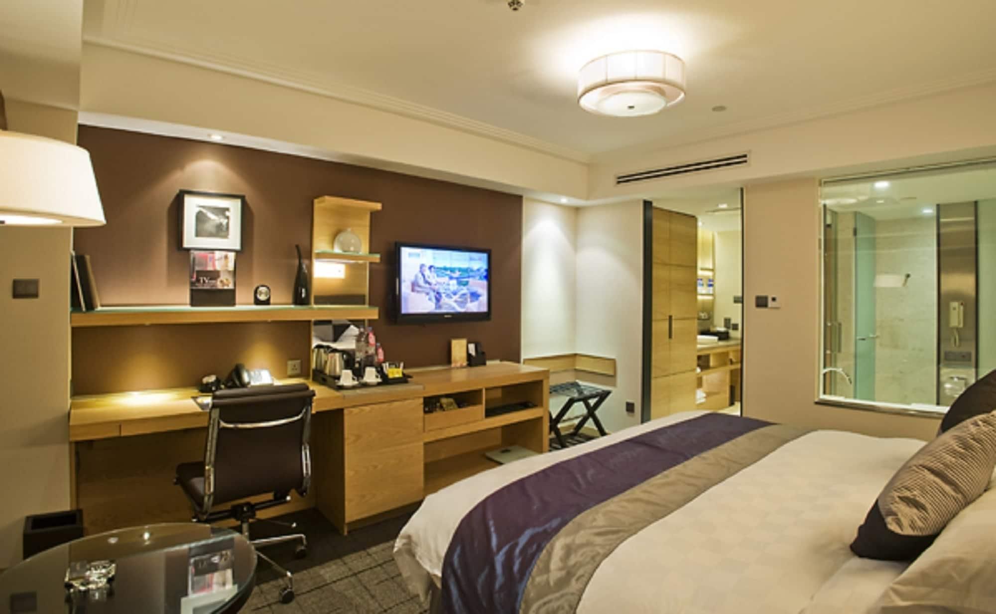 A hotel room with a bed, desk, chair, television, and a bathroom visible through a glass door.