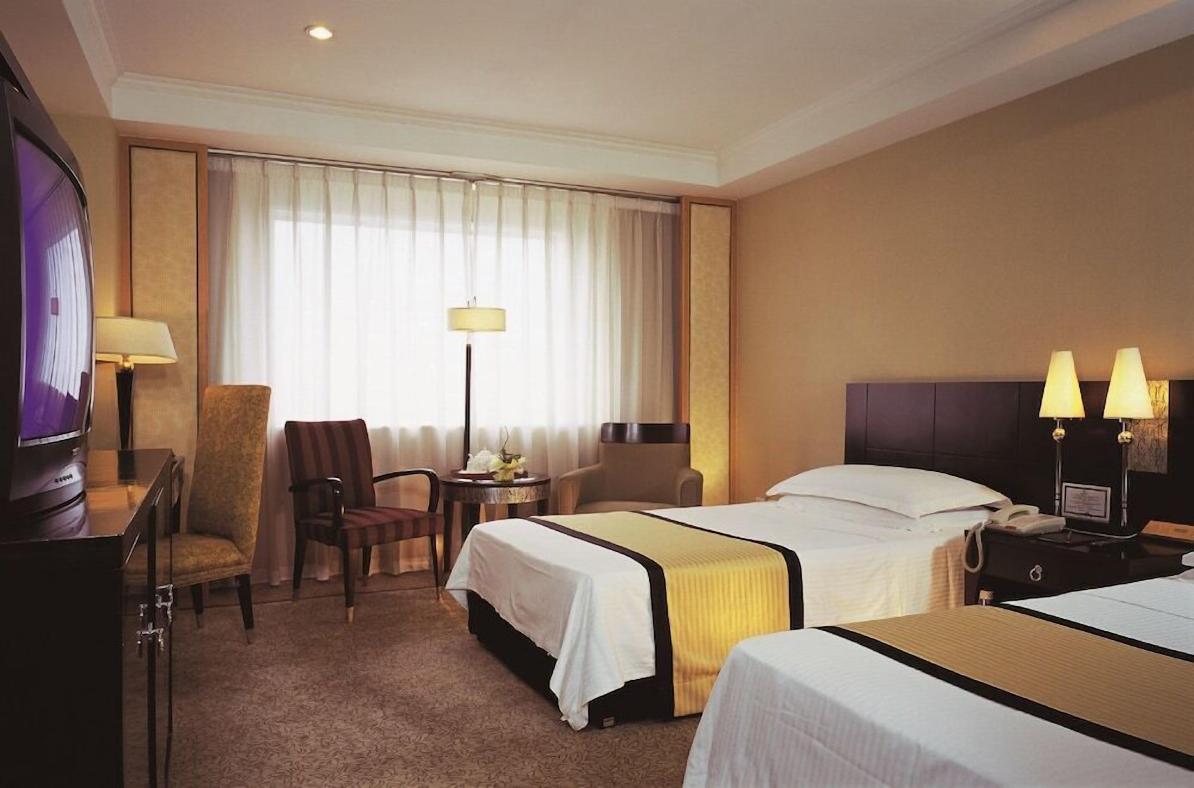 Habitación de hotel con dos camas, un escritorio con televisor, una silla, una lámpara y un ventanal con cortinas.