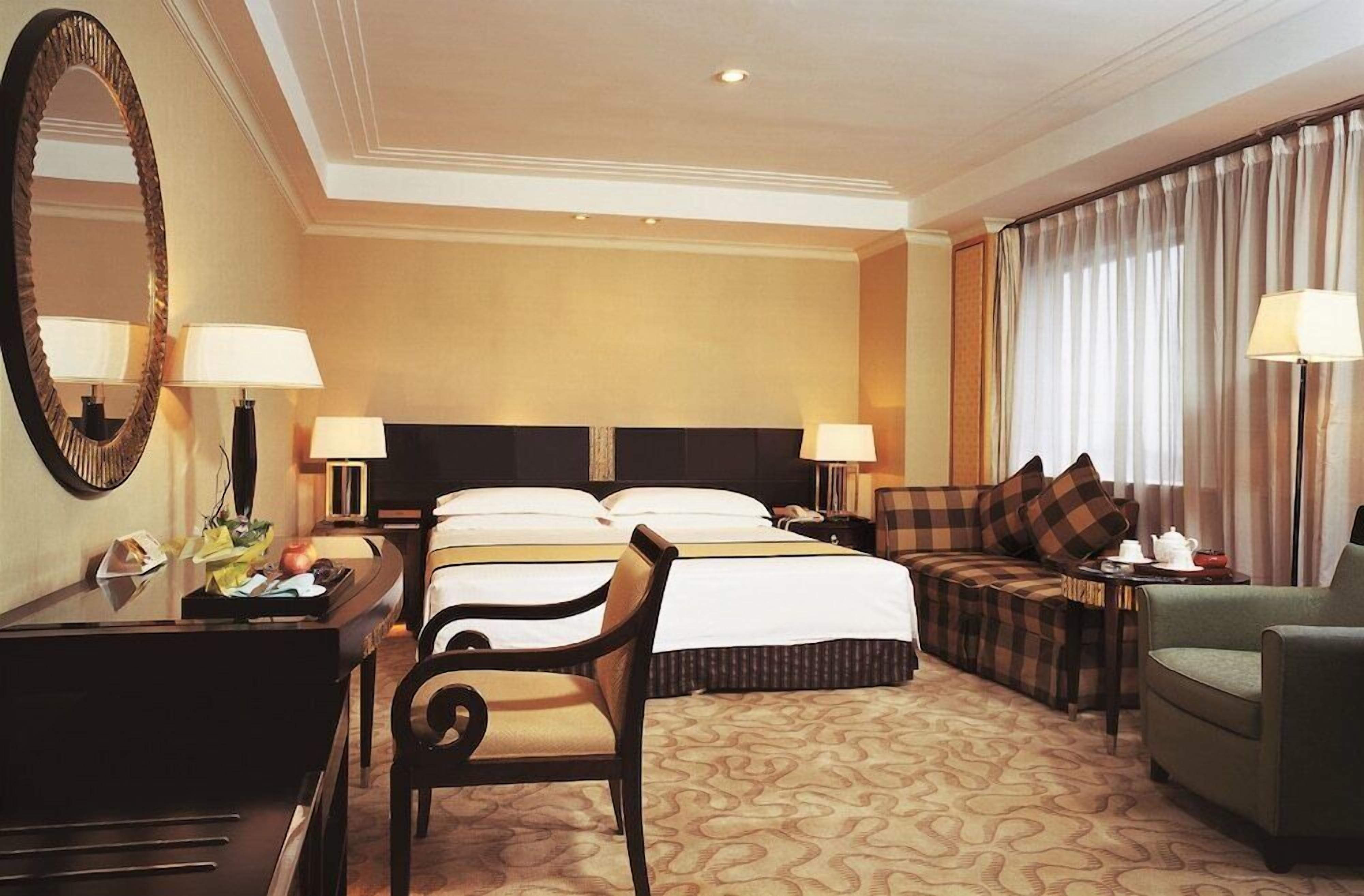 Une chambre d’hôtel avec un grand lit, deux tables de chevet avec des lampes, un bureau avec une chaise, un canapé et un grand miroir.