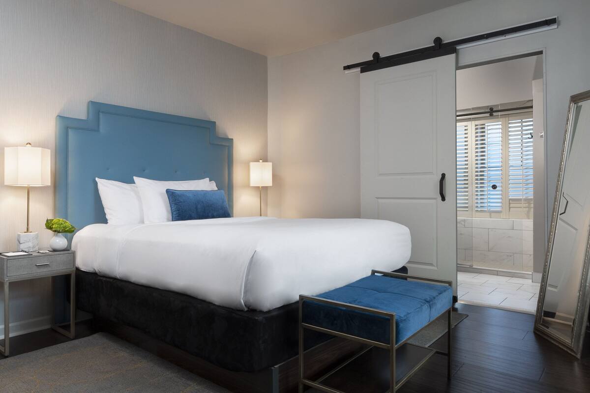 A modern bedroom with a large bed, bedside lamps, a nightstand, and a view of the outside through a sliding door.