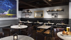 A restaurant interior with round tables, dark wood chairs, a black leather booth, and a large painting on the wall. - Seven Gables, St. Louis West, a Tribute Portfolio Hotel (St. Louis)