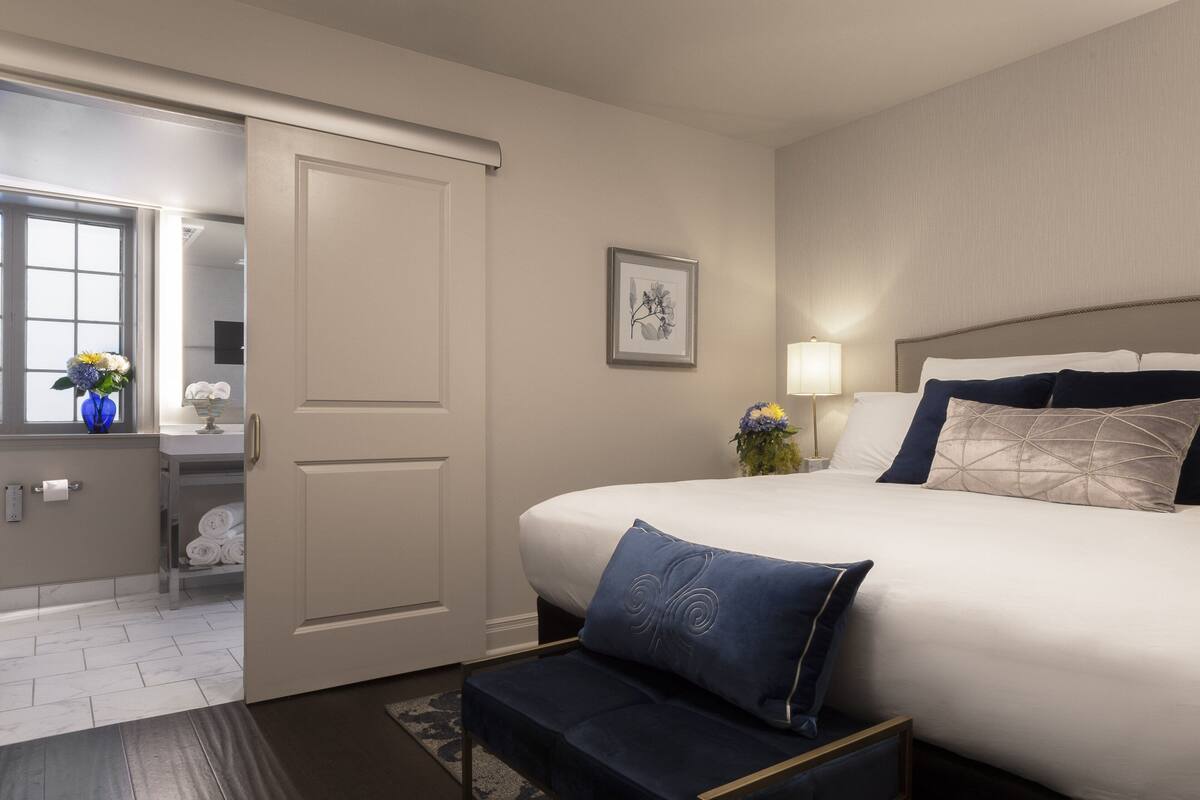 A hotel room with a large bed, a bedside table with a lamp, a framed picture on the wall, and a bathroom visible through an open door.