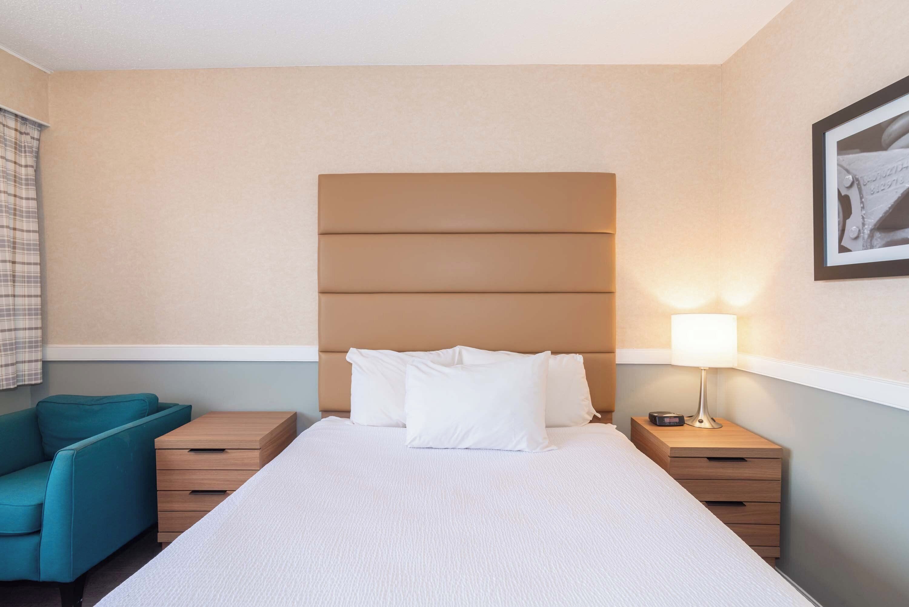 A hotel room with a large bed, a nightstand with a lamp, a wooden dresser, and a blue armchair.