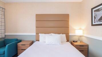 A hotel room with a large bed, a nightstand with a lamp, a wooden dresser, and a blue armchair.