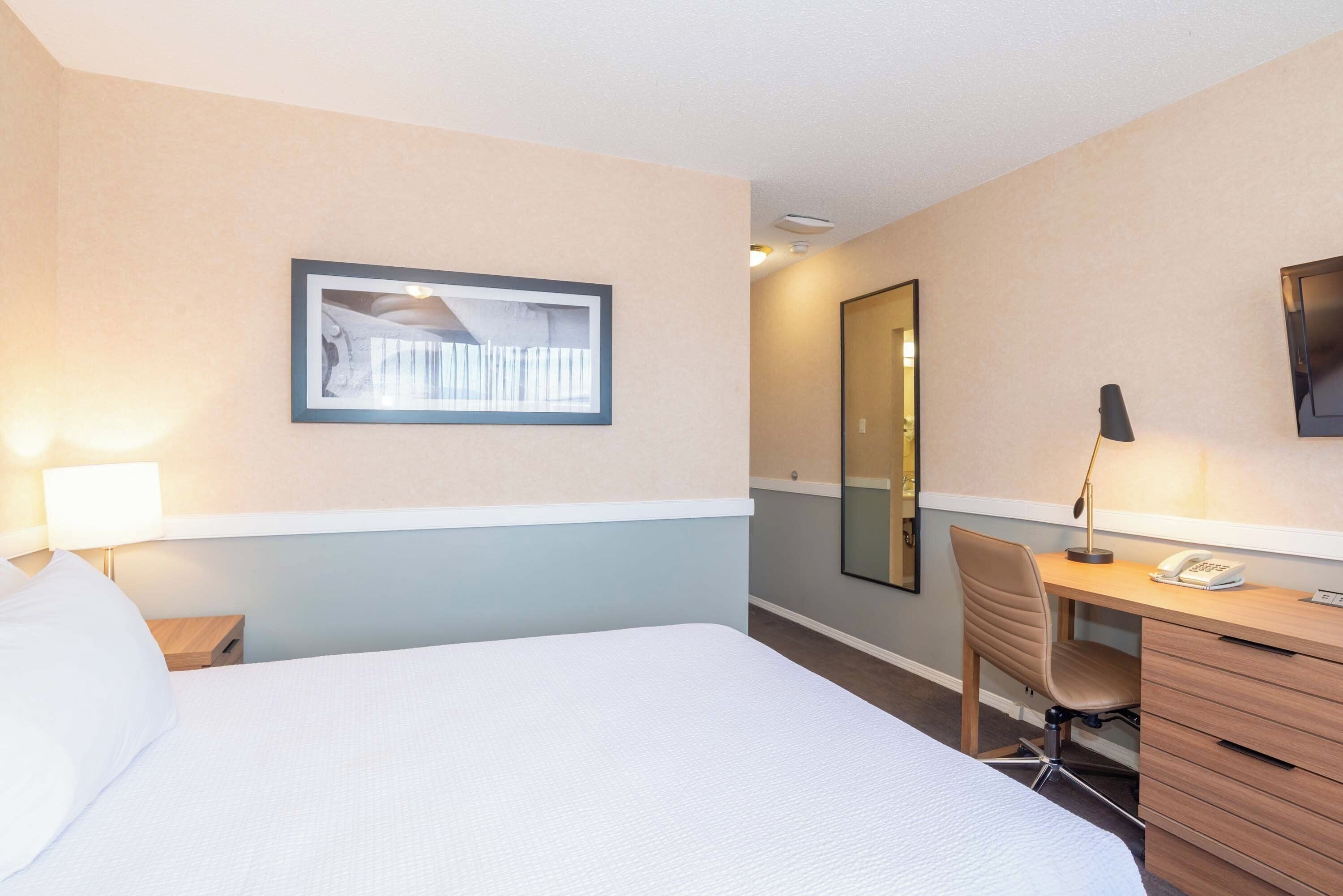 Habitación de hotel con cama, escritorio, silla, lámpara, teléfono y un cuadro en la pared.
