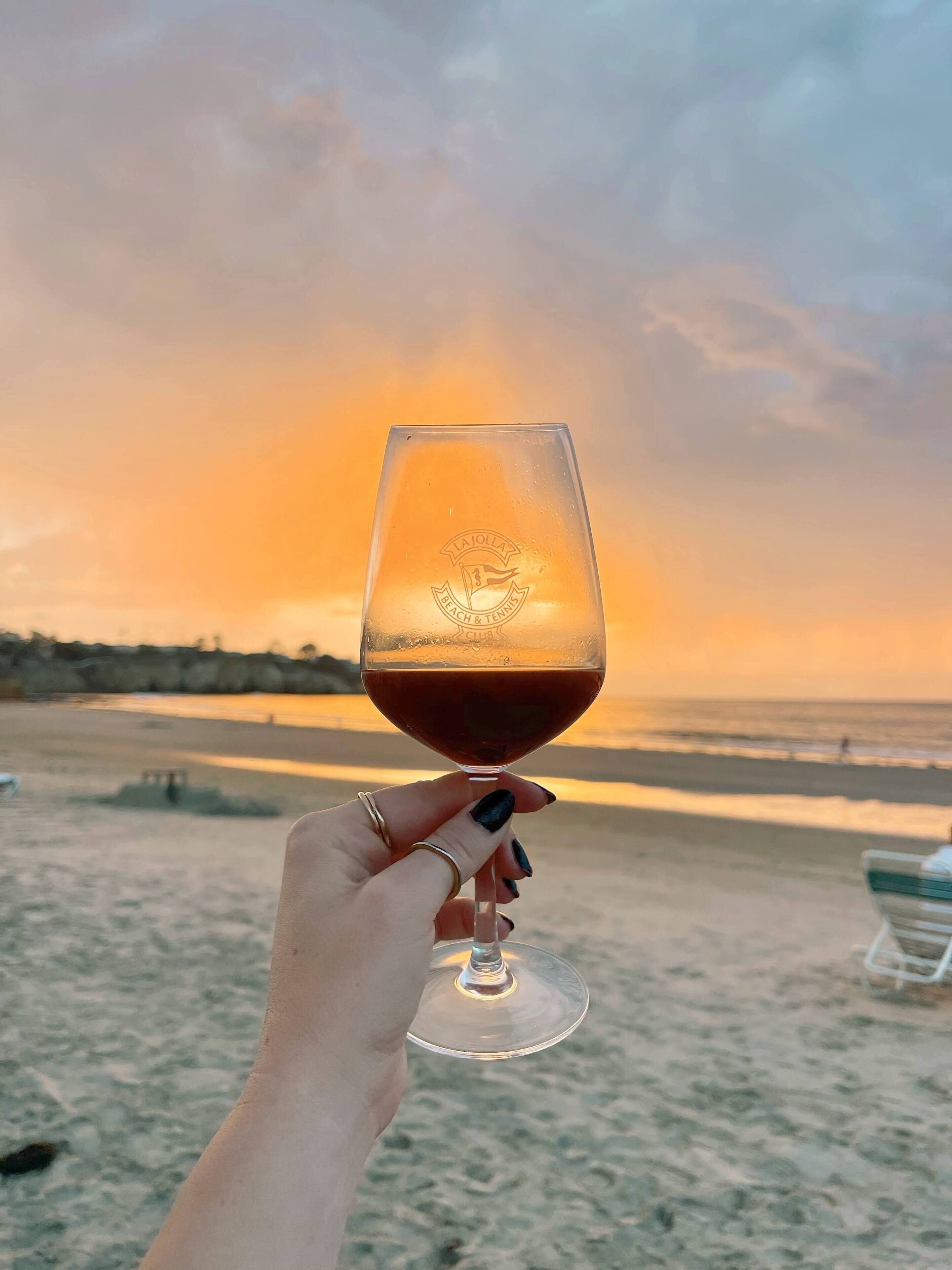 A hand holding a wine glass with a beach and sunset in the background.