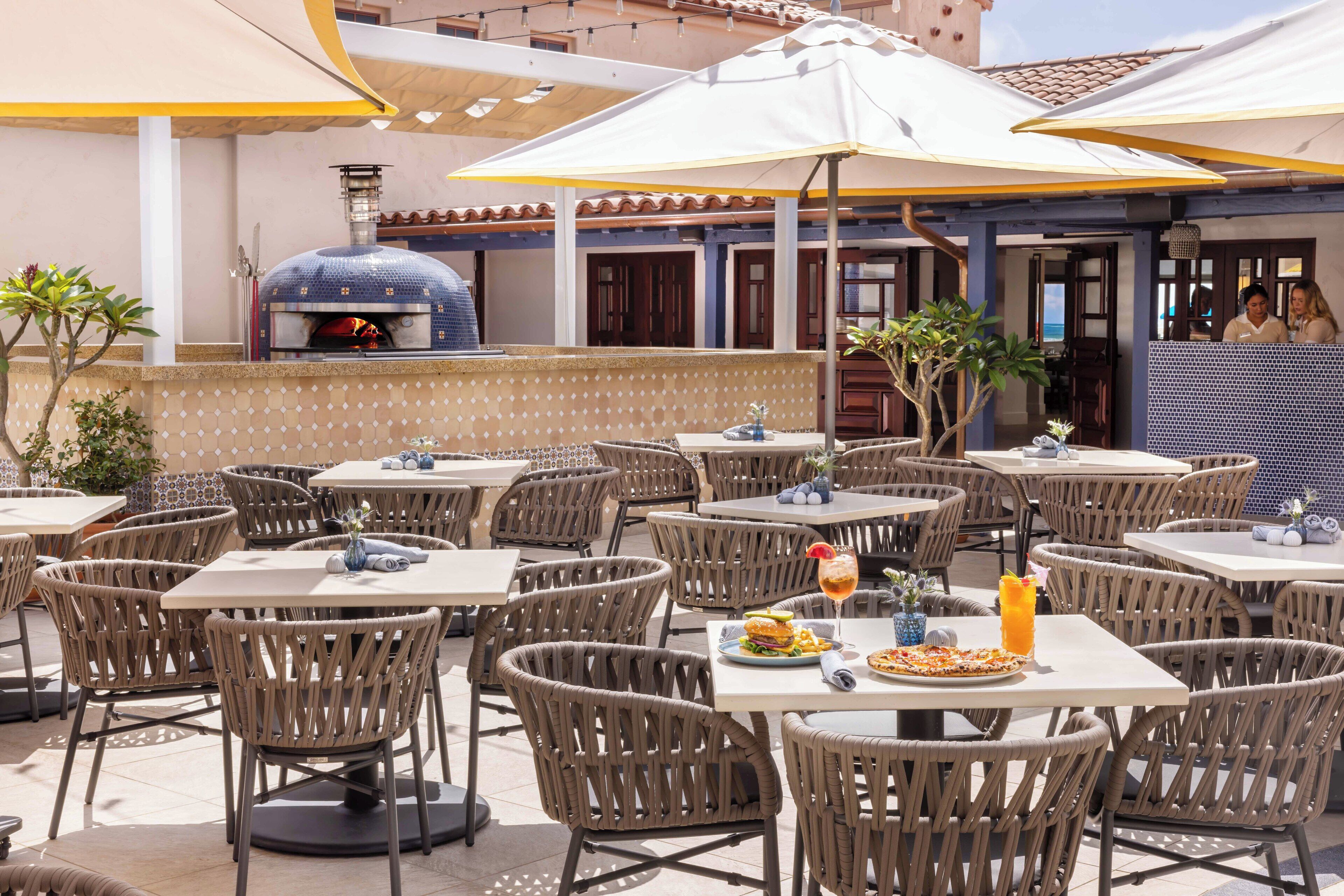 An outdoor dining area with wicker chairs and tables, a pizza oven, and umbrellas providing shade.
