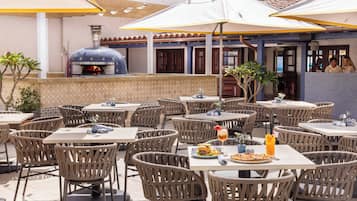 An outdoor dining area with wicker chairs and tables, a pizza oven, and umbrellas providing shade.