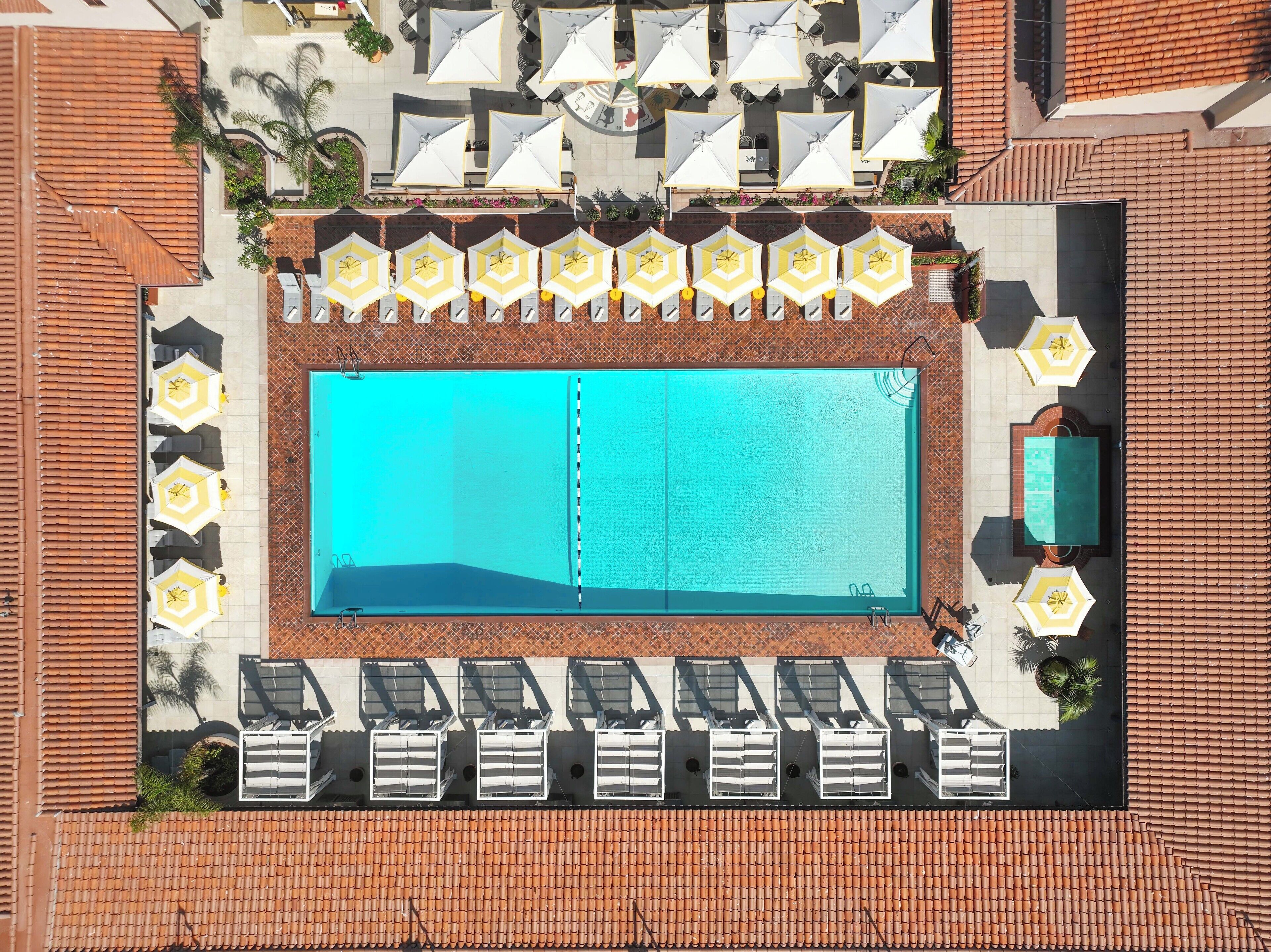 Ein Luftbild eines Hotels mit einem rechteckigen Pool, umgeben von Liegestühlen und Sonnenschirmen, und einem gepflasterten Dach.