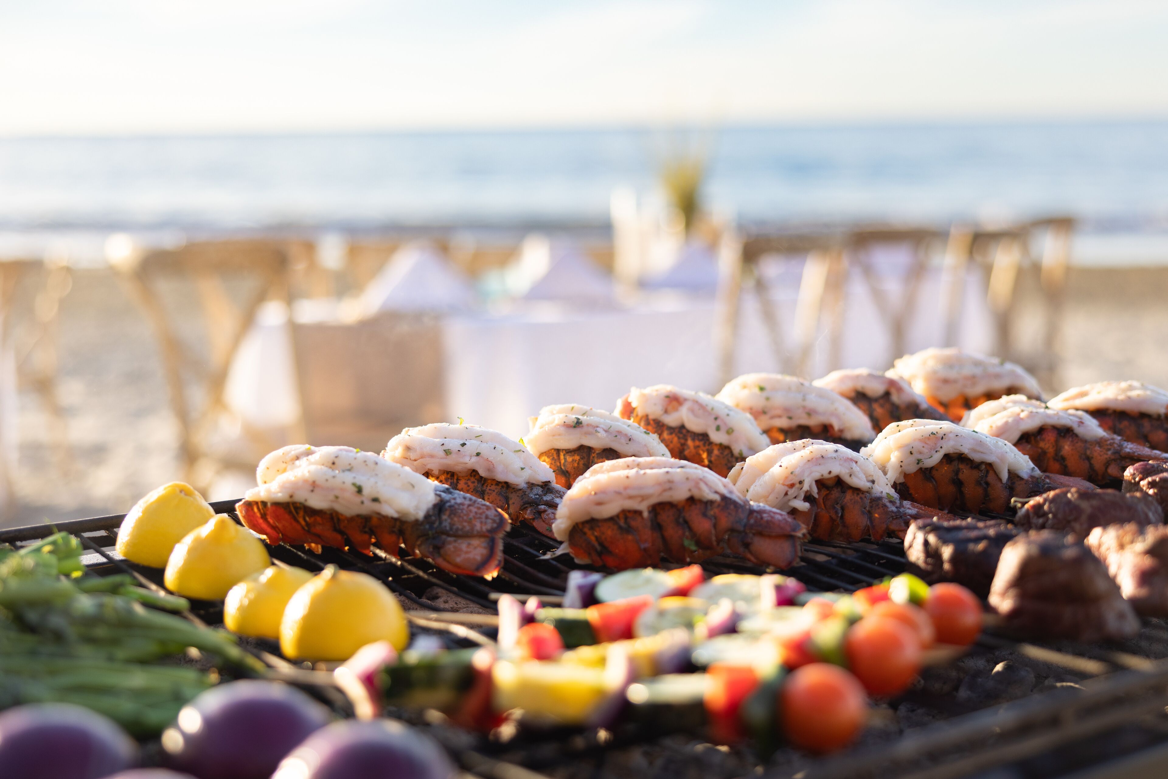 Un barbecue avec des légumes et des fruits de mer grillés, installé sur une plage avec vue sur l’océan.