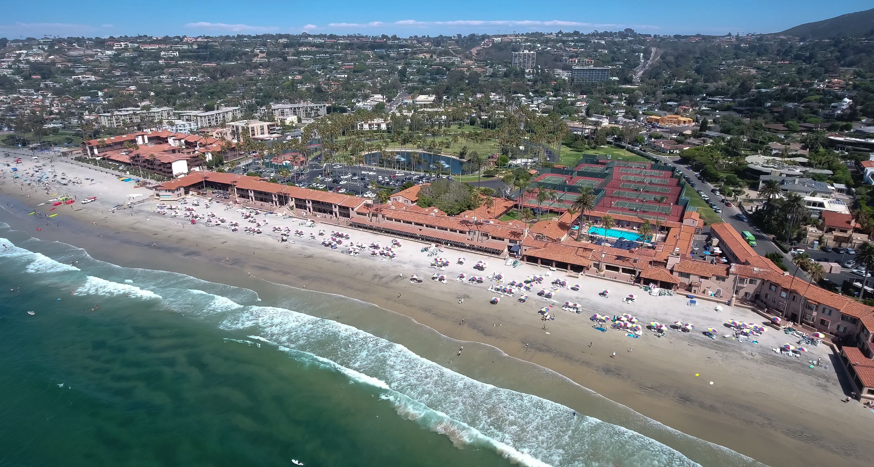 An aerial view of a coastal area with a beach, a parking lot, and a sports complex.