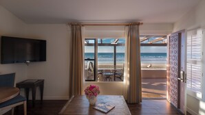 A room with a TV, a wooden table with a book and a vase of flowers, a window with beach view, and a door with patterned design. - La Jolla Beach and Tennis Club (La Jolla)