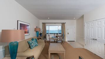 A hotel room with a sofa, a coffee table, a dining table with chairs, a view of the sea through a window, and a door.