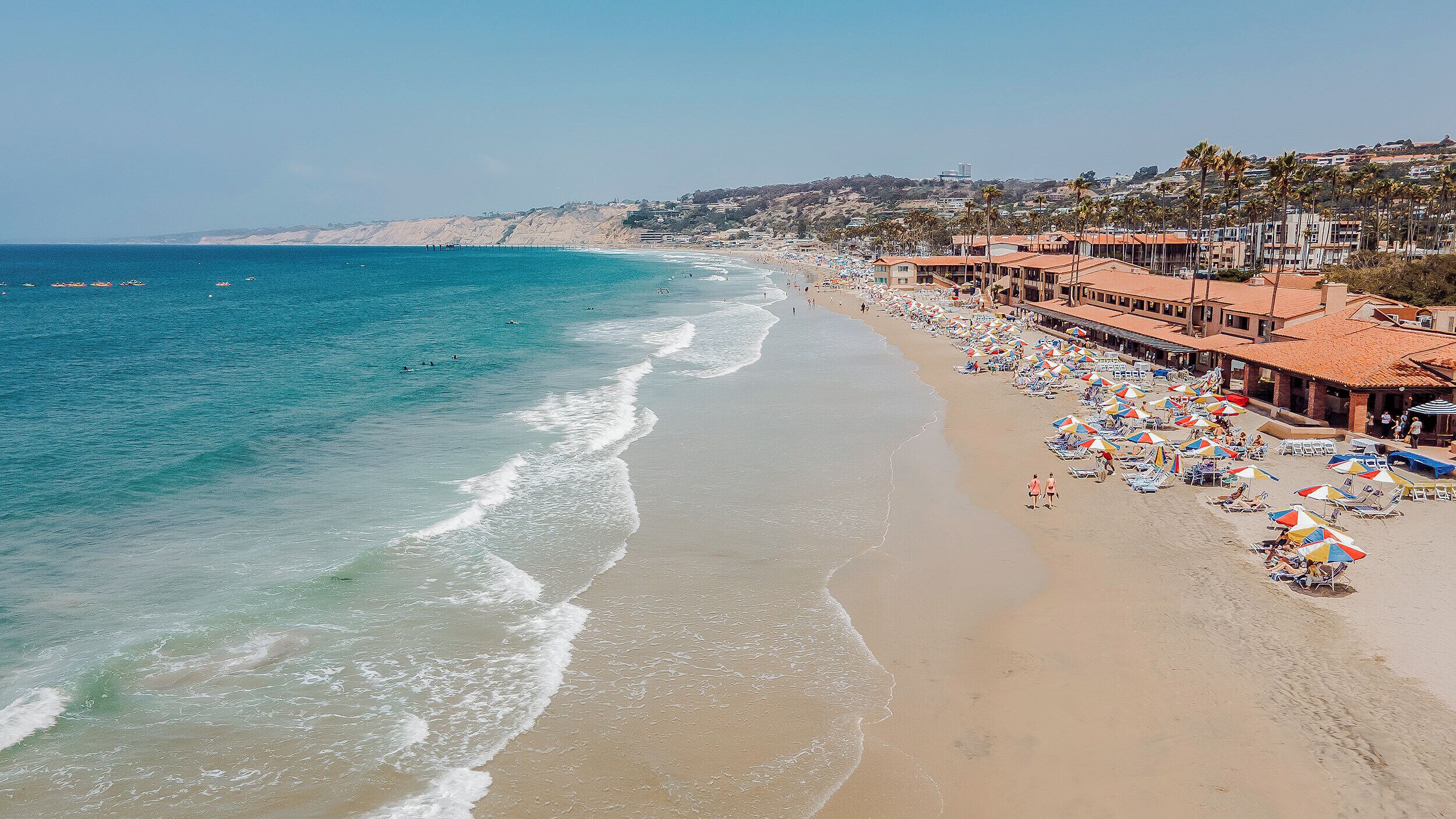 A beach with a long stretch of sand, a row of beach umbrellas, and a few people walking.