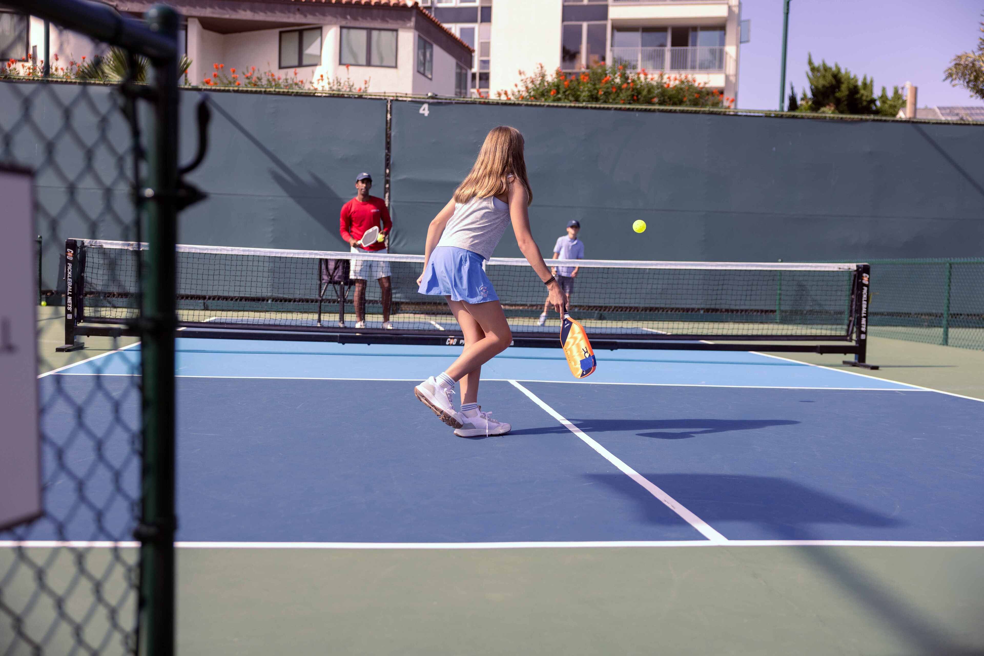 Una cancha de tenis con red, una jugadora con falda azul y remera blanca, y otra con remera roja.