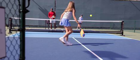 Una cancha de tenis con red, una jugadora con falda azul y remera blanca, y otra con remera roja.