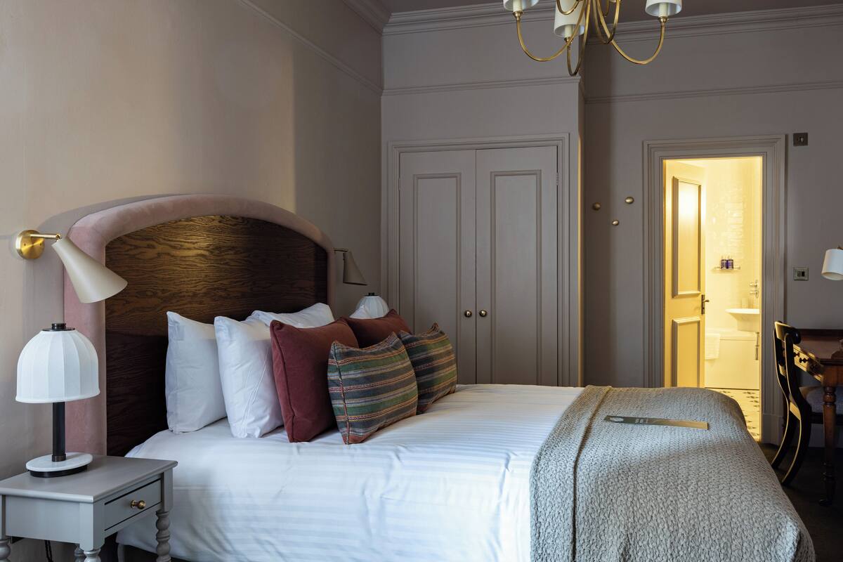 A neatly arranged bedroom with a large bed, a bedside table with a lamp, and a door leading to a bathroom.