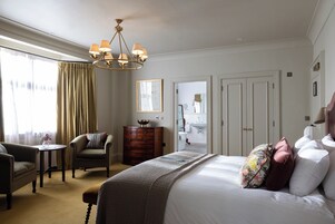 Una habitación de hotel con una cama grande, una zona de estar con una silla y una mesa, un aparador y una araña.