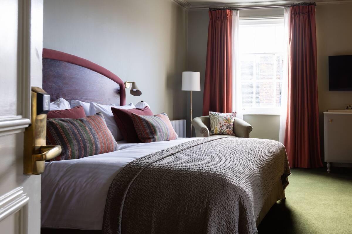 A neatly made bed with a red headboard, a bedside lamp, and a chair with a floral cushion.