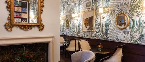 A room with a fireplace, a mirror, and a patterned wallpaper. There are chairs and a table with a vase of flowers.