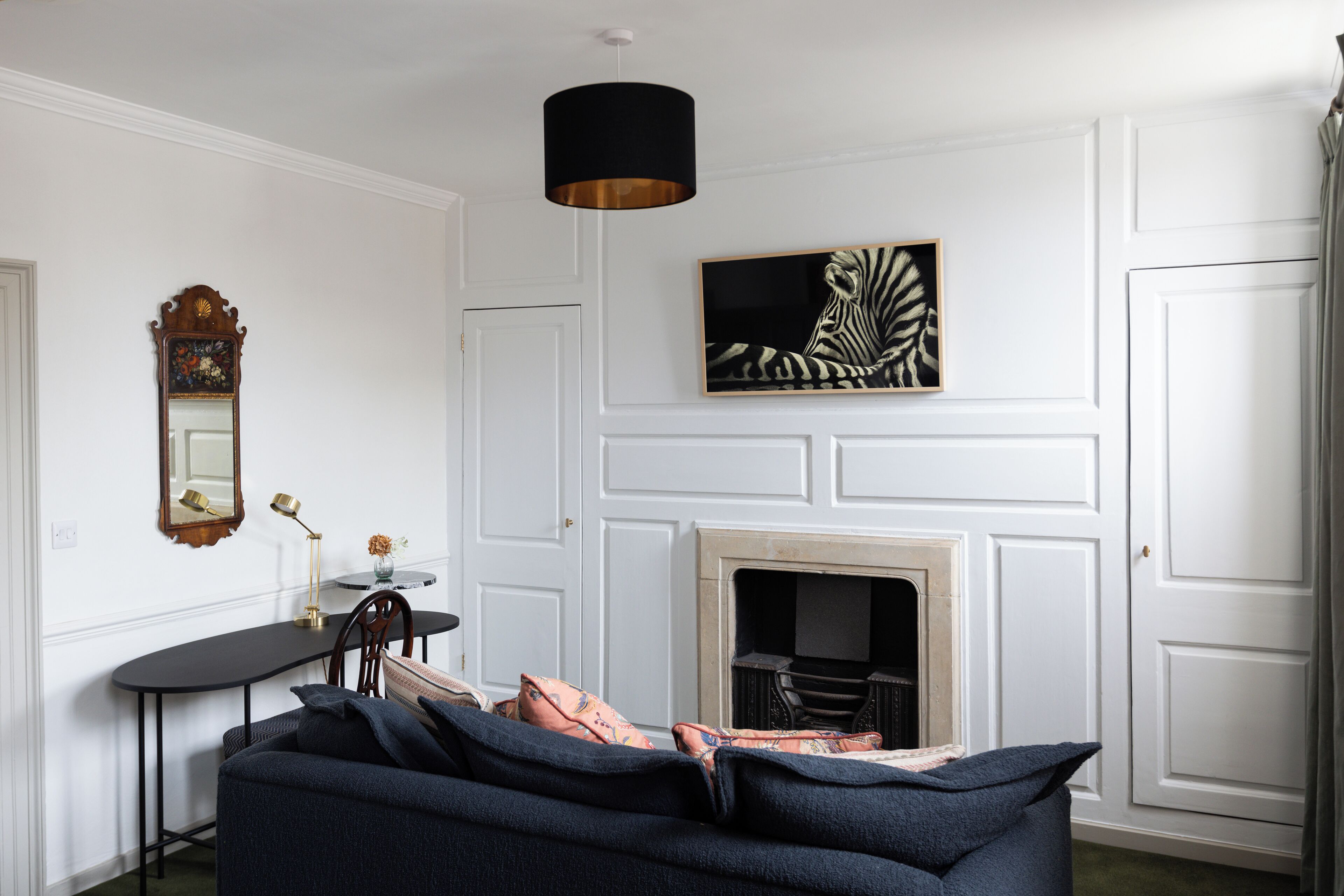 A room with a dark blue sofa, a black lamp, a framed zebra painting, a fireplace, and a wooden mirror.