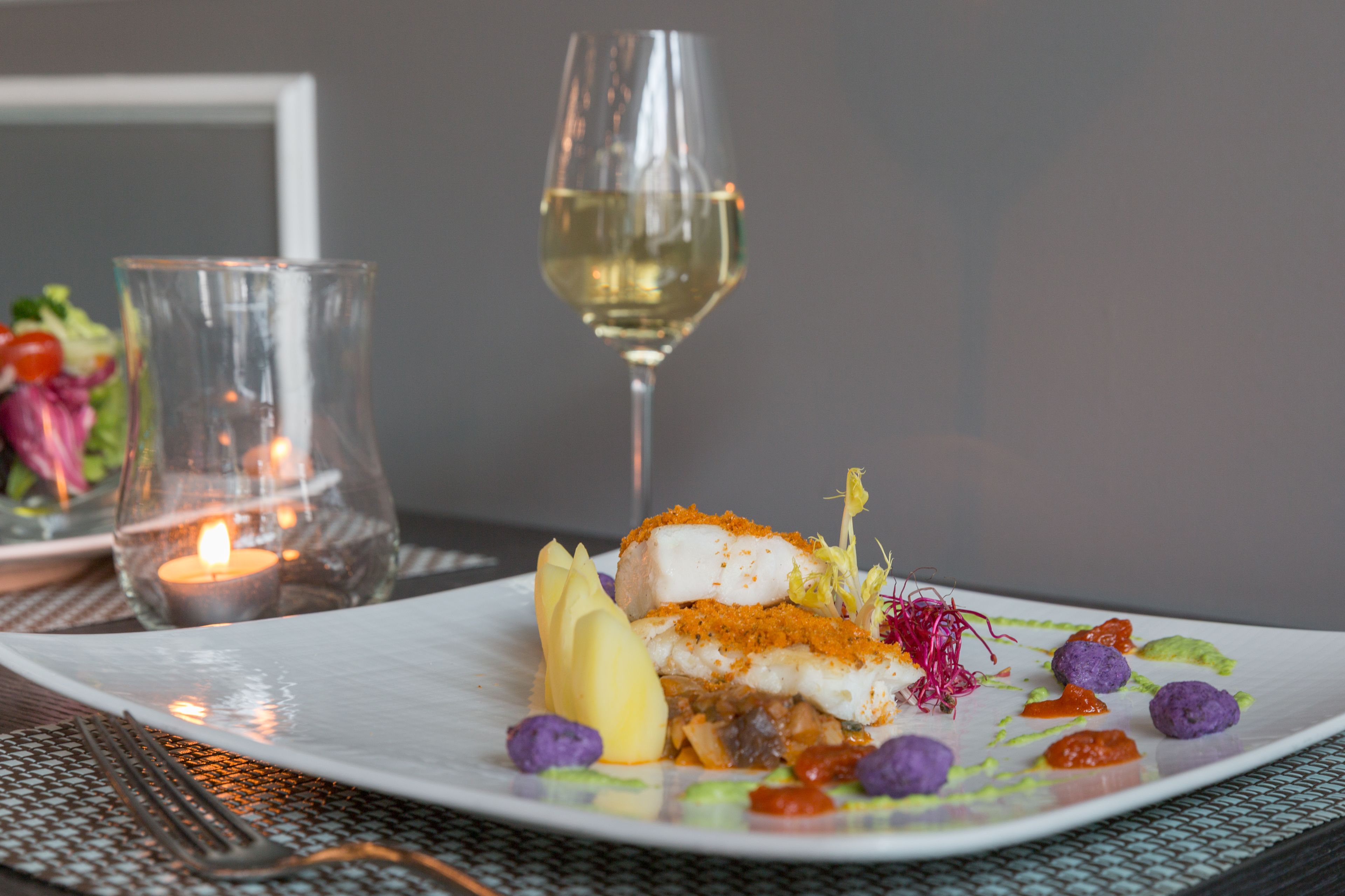 Un plato blanco con un guiso de pescado, una copa de vino blanco y una vela sobre una mesa.