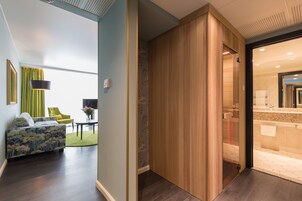 Et moderne hotelværelse med en træafskærmning, et opholdsområde med sofa og stole, samt et glimt af et badeværelse med vask og spejl.