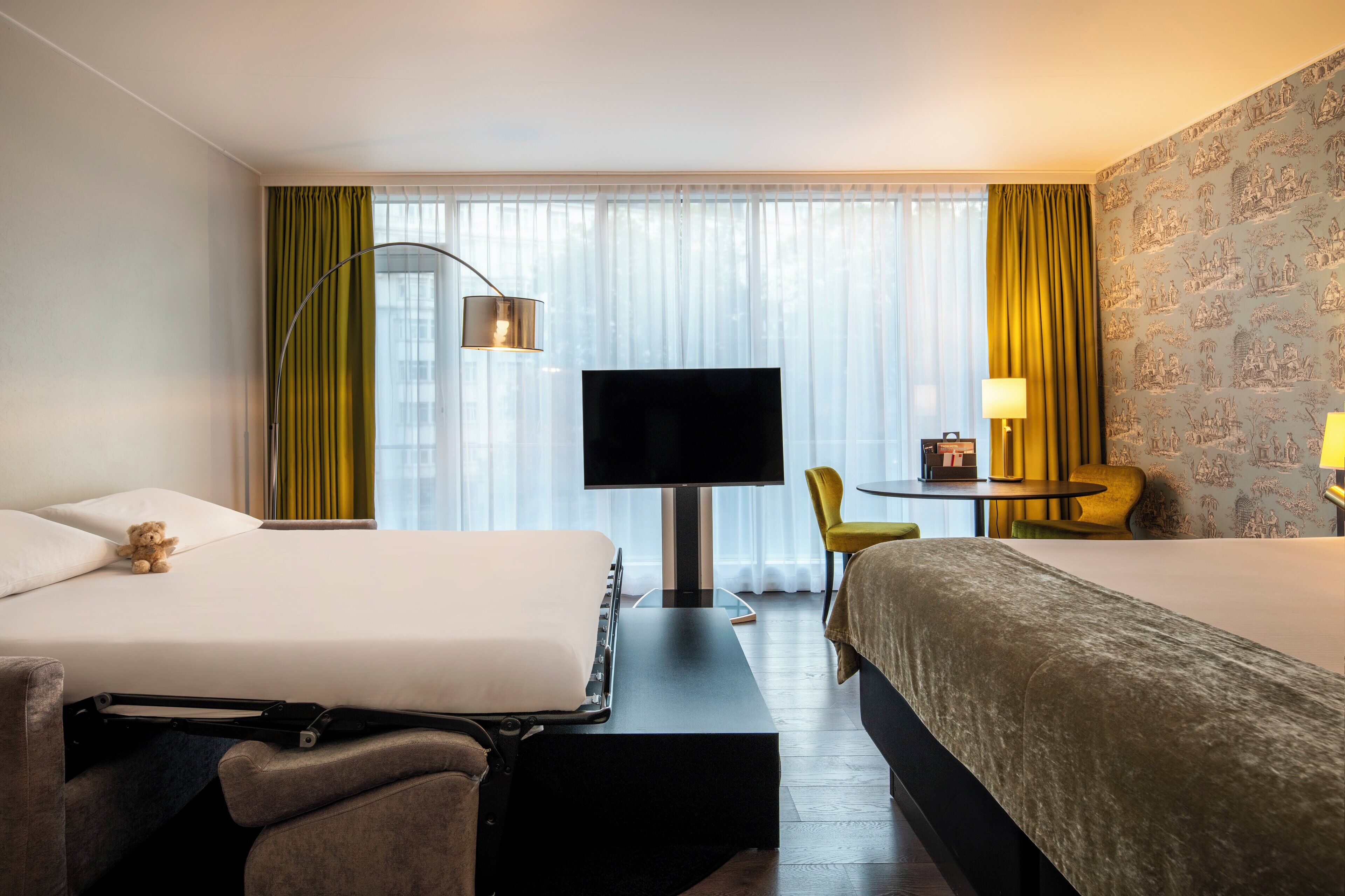 Ein Hotelzimmer mit zwei Betten, einem Fernseher, einem Schreibtisch mit Stuhl und einem kleinen Tisch mit Lampe.