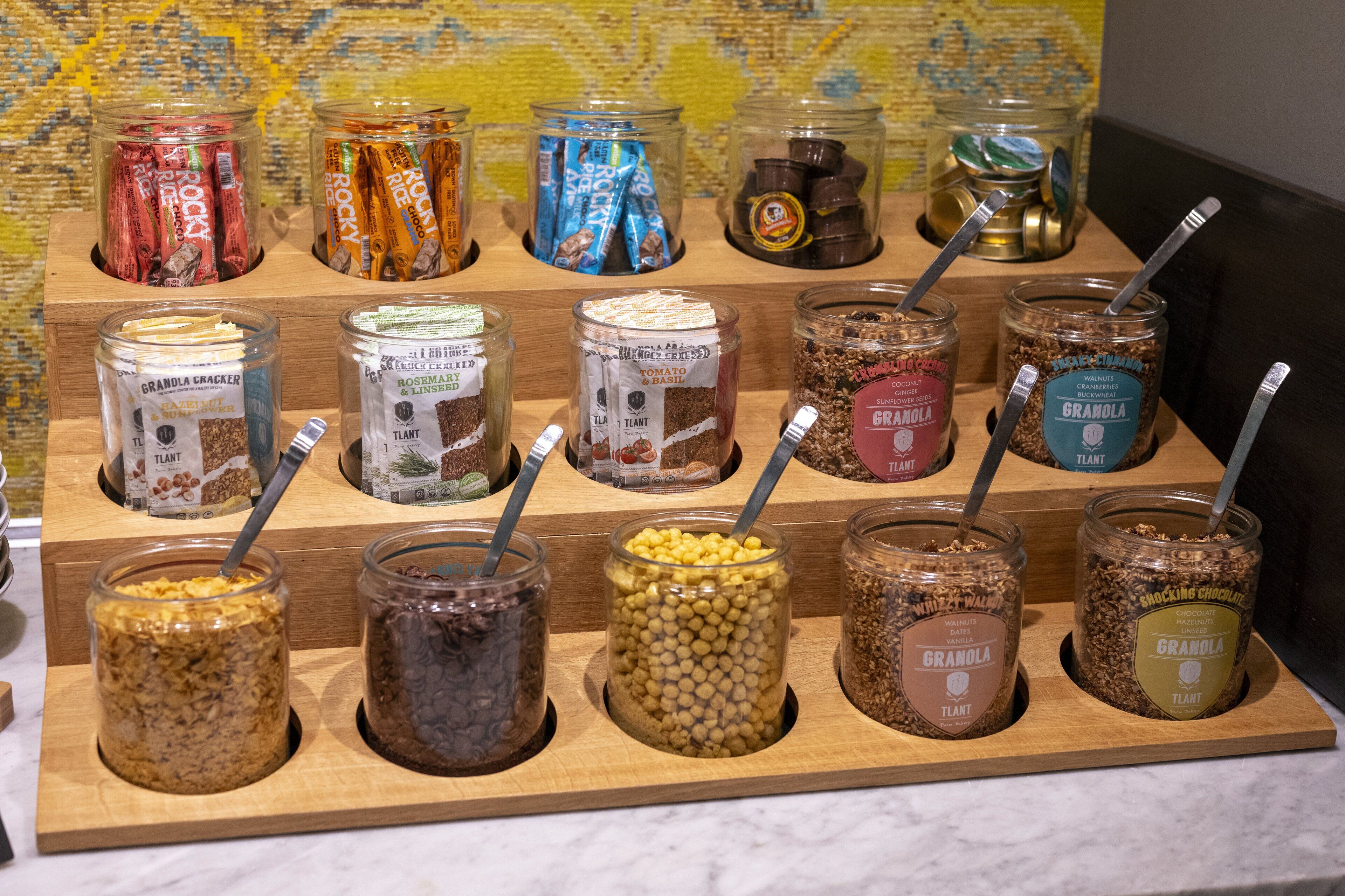 A display of various granola and cereal bars in glass jars with labels, arranged on a wooden board.