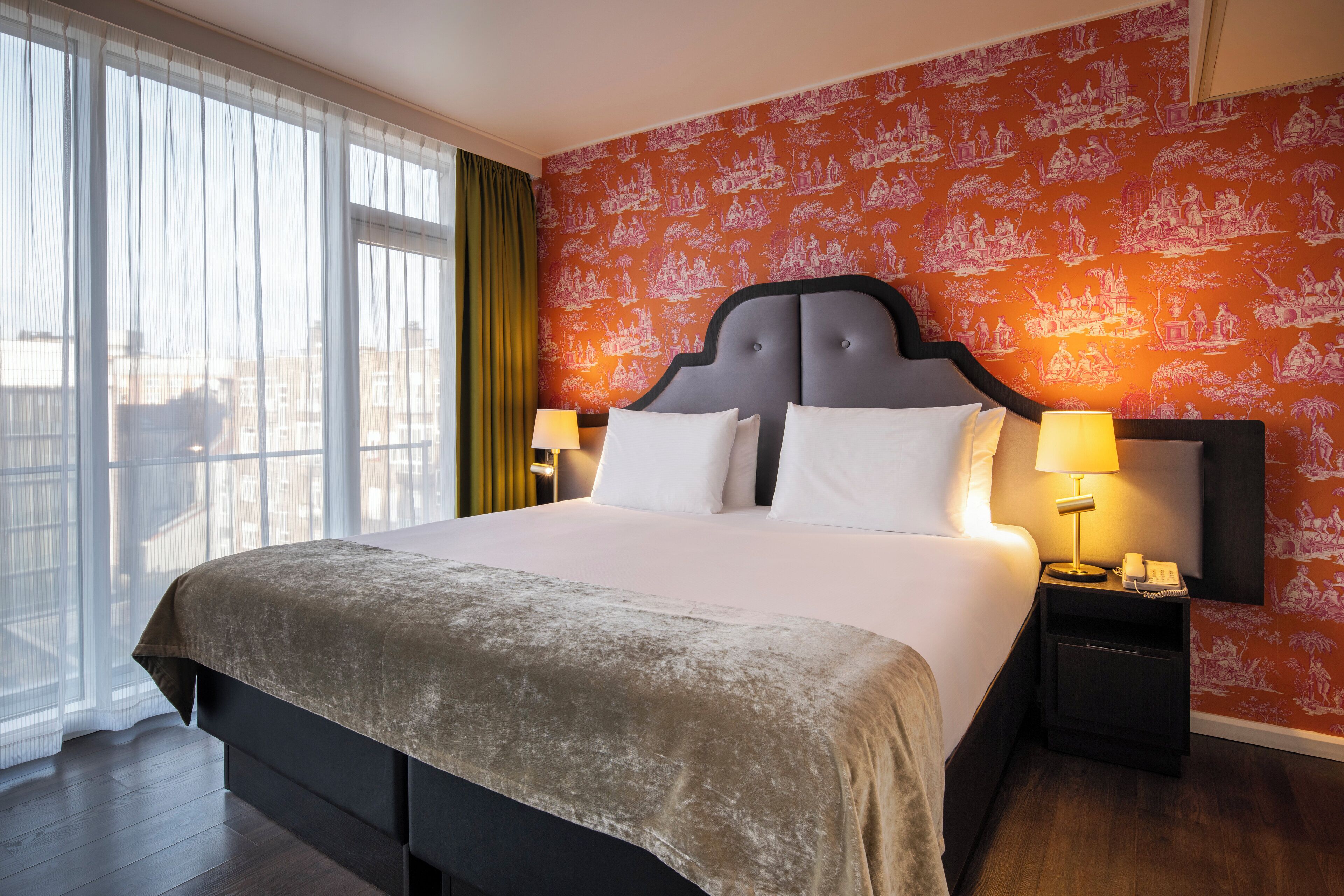 A hotel room with a large bed, two bedside tables, a nightstand, a window with sheer curtains, and a patterned wallpaper.