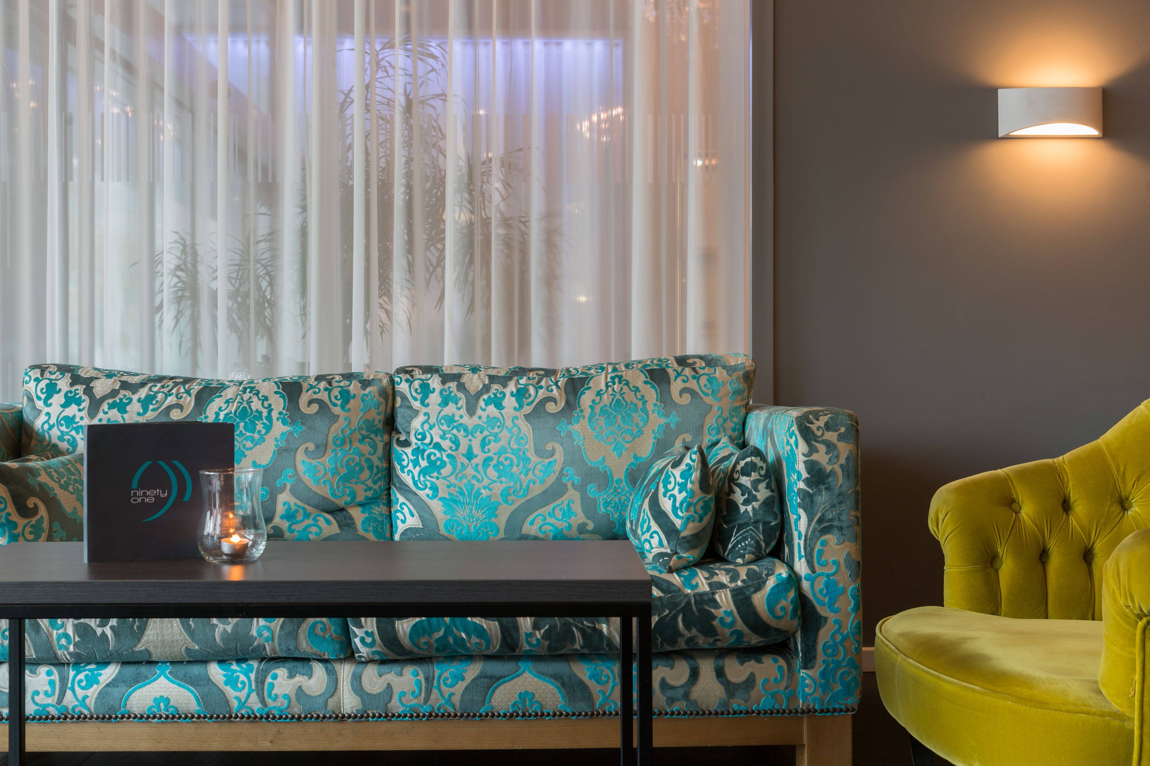 A room with a patterned sofa, a small table with a glass and a black box, a yellow armchair, and a wall-mounted light fixture.