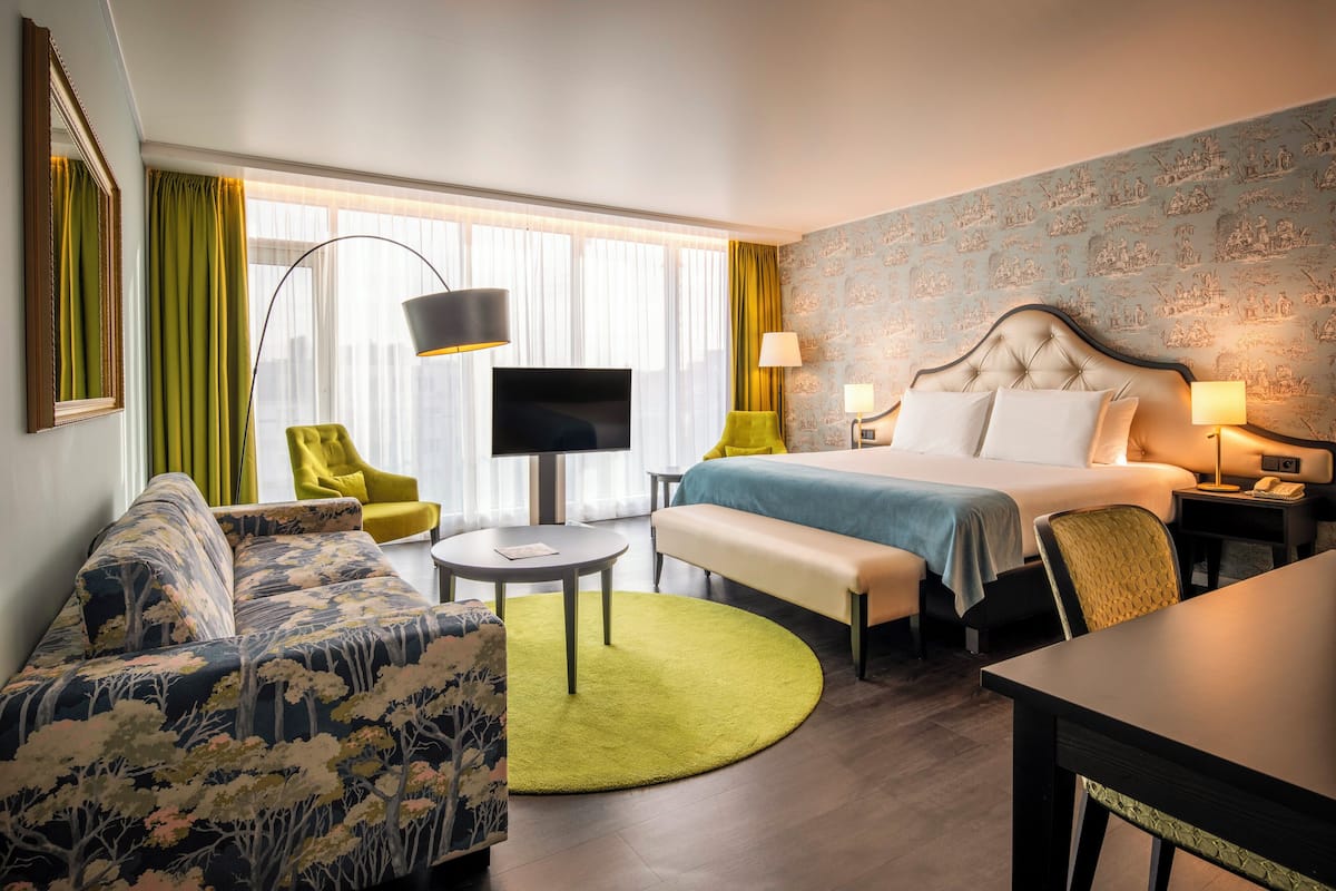 A hotel room with a bed, a sofa, a desk, a chair, a TV, and a lamp.