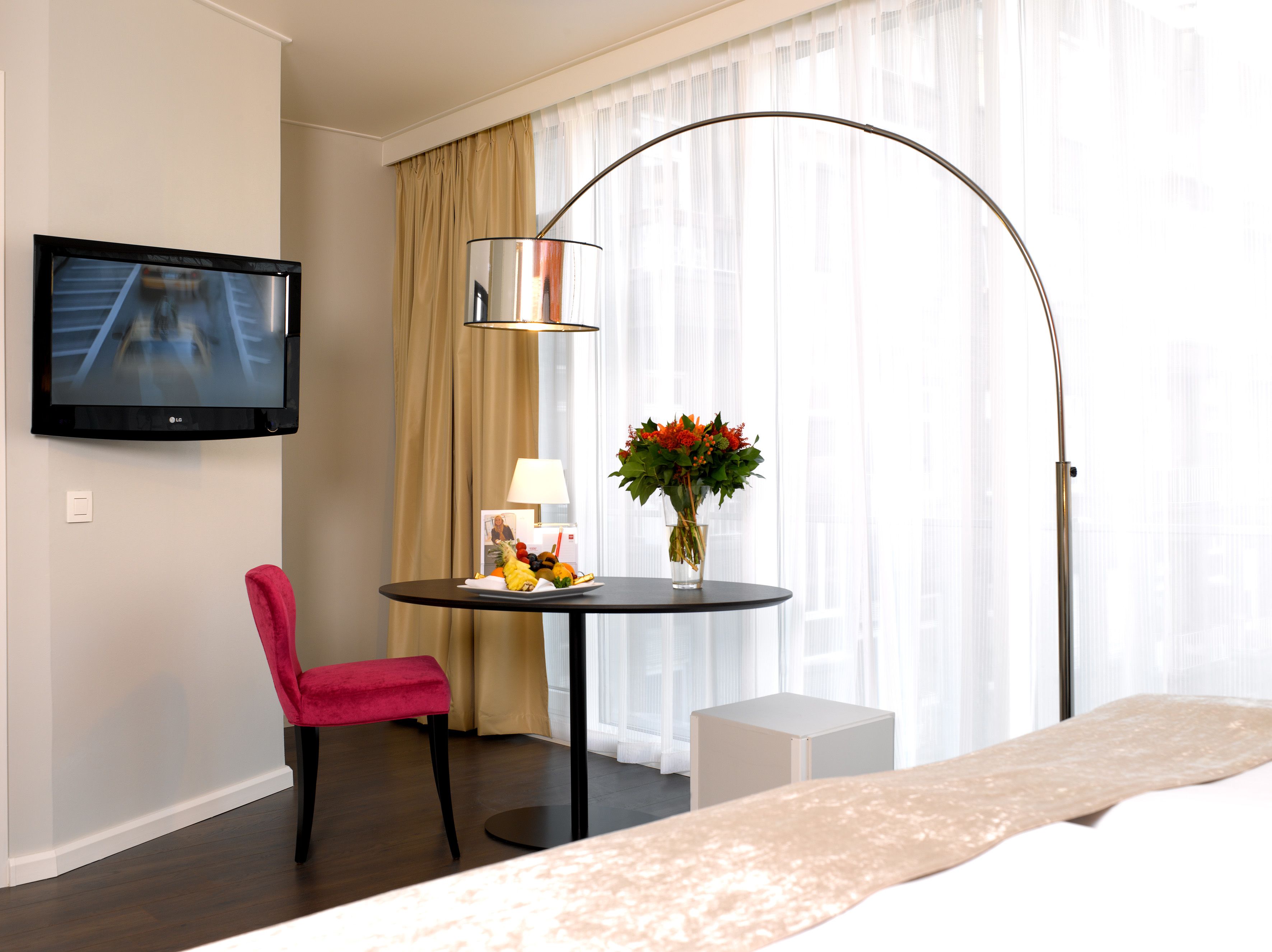 A hotel room with a bed, a TV mounted on the wall, a round table with a lamp and a vase of flowers, a red chair, and a small white side table.