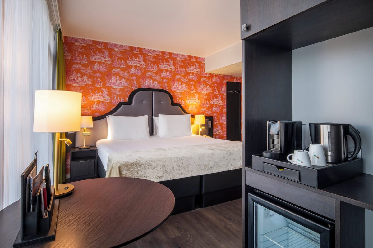 A hotel room with a bed, bedside lamps, a coffee maker, a mini-fridge, and a desk with a lamp.