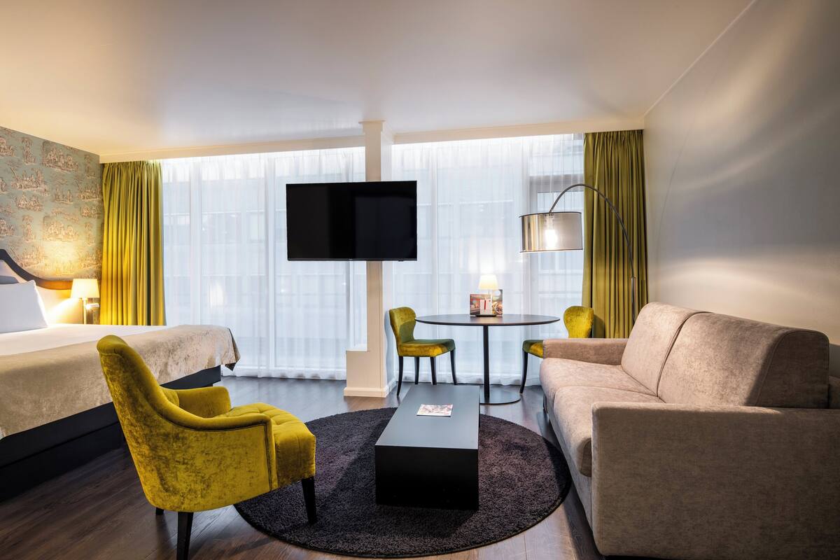 A hotel room with a bed, two armchairs, a sofa, a coffee table, a TV, and a lamp.