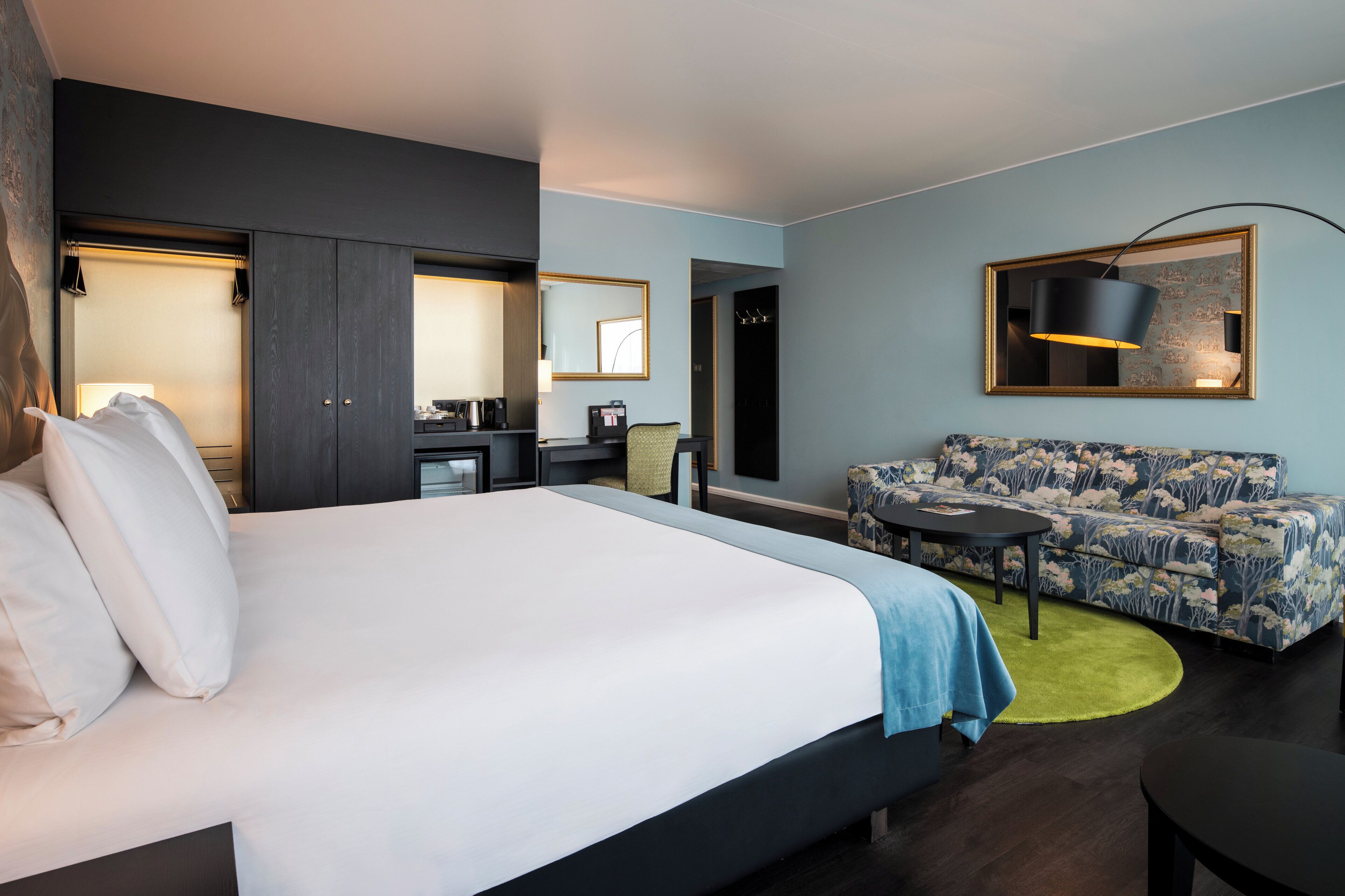 Une chambre d’hôtel moderne dotée d’un grand lit, d’un canapé, d’un bureau avec une chaise, d’une machine à café et d’un miroir.