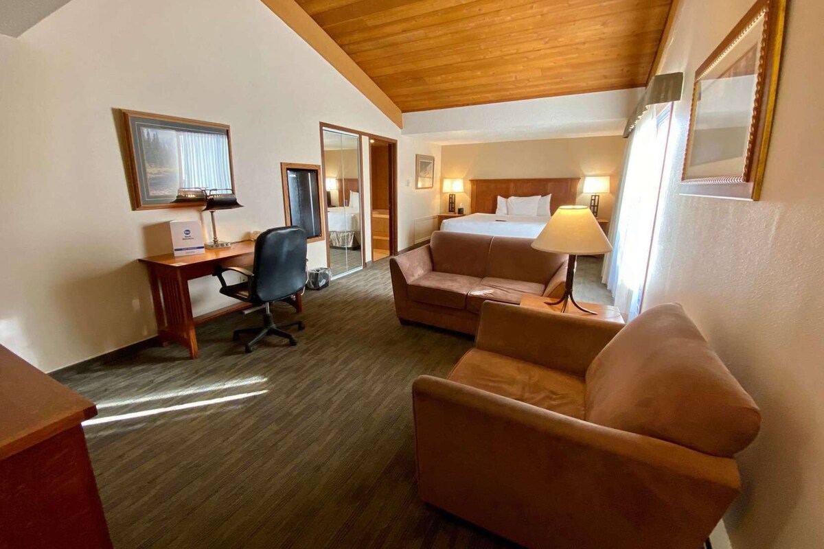 A hotel room with a wooden ceiling, a desk with a chair, a bed with a headboard, a sofa, a lamp, and a mirror.