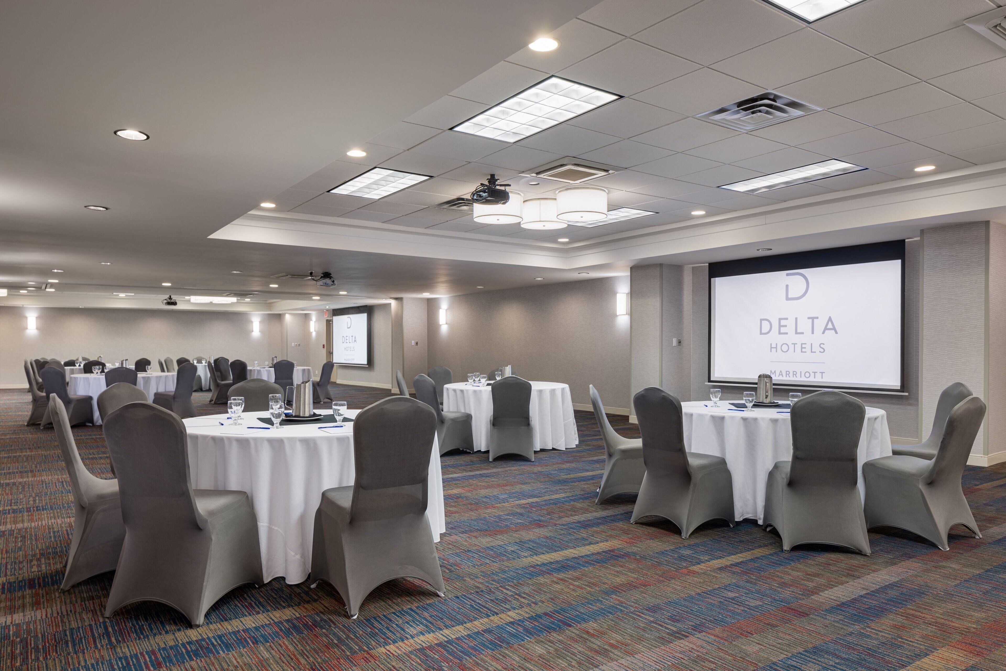 A conference room with round tables, gray chairs, and a projector screen displaying 'Delta Hotels Marriott'.