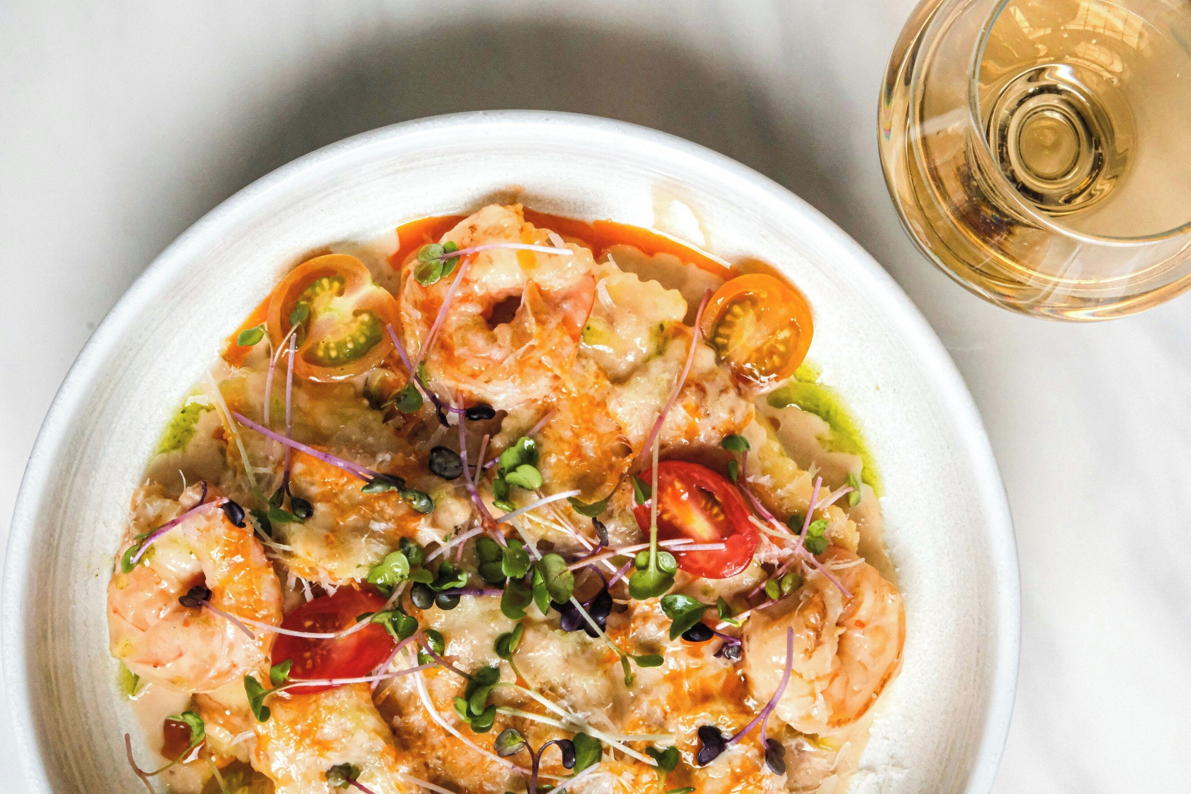 Eine Schüssel mit Meeresfrüchtsalat, darunter Garnelen, Tomaten und Sprossen, dazu ein Glas Weißwein.