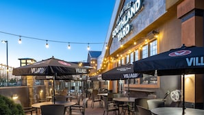 An outdoor patio area with multiple tables and chairs, covered by umbrellas with the 'VILLAGE' logo, and a building with 'SOUTHLAND YARD' signage. - Delta Hotels by Marriott Calgary South (Calgary)