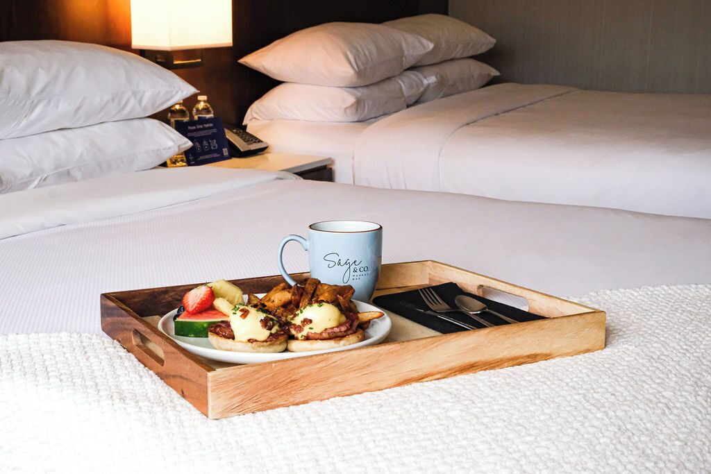 A hotel room with a neatly made bed, a tray with breakfast including eggs, fruit, and bacon, and a mug labeled "Sage & Co."