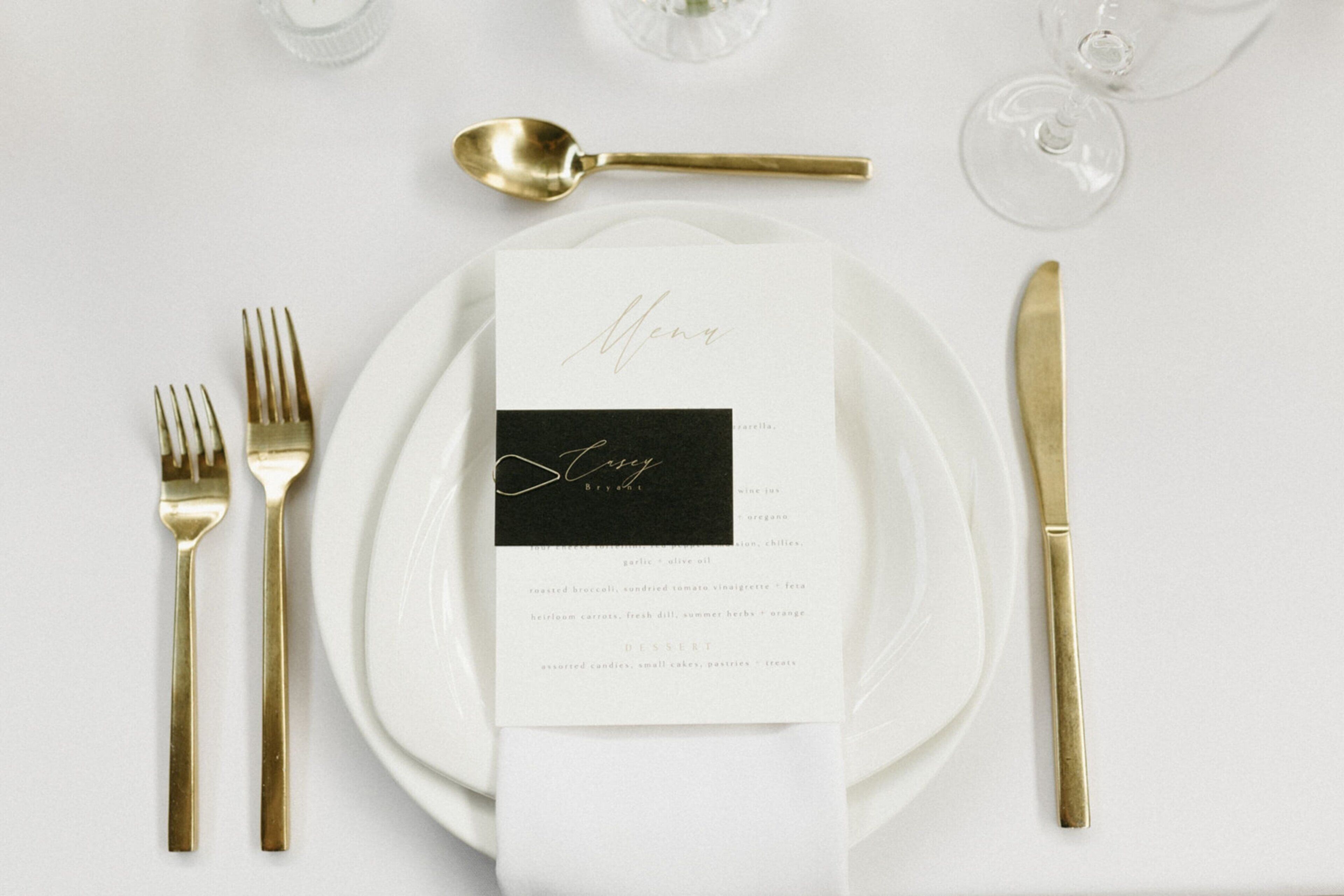 A table setting with a menu, gold cutlery, and wine glasses.